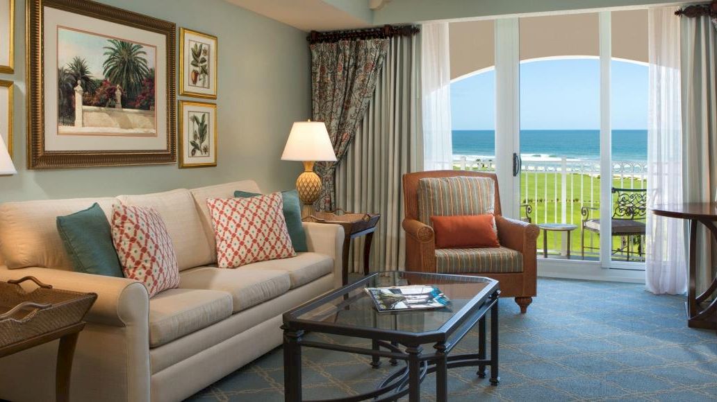A cozy living room with a sofa, armchair, lamps, and framed art faces a balcony overlooking the sea, offering a relaxing view.