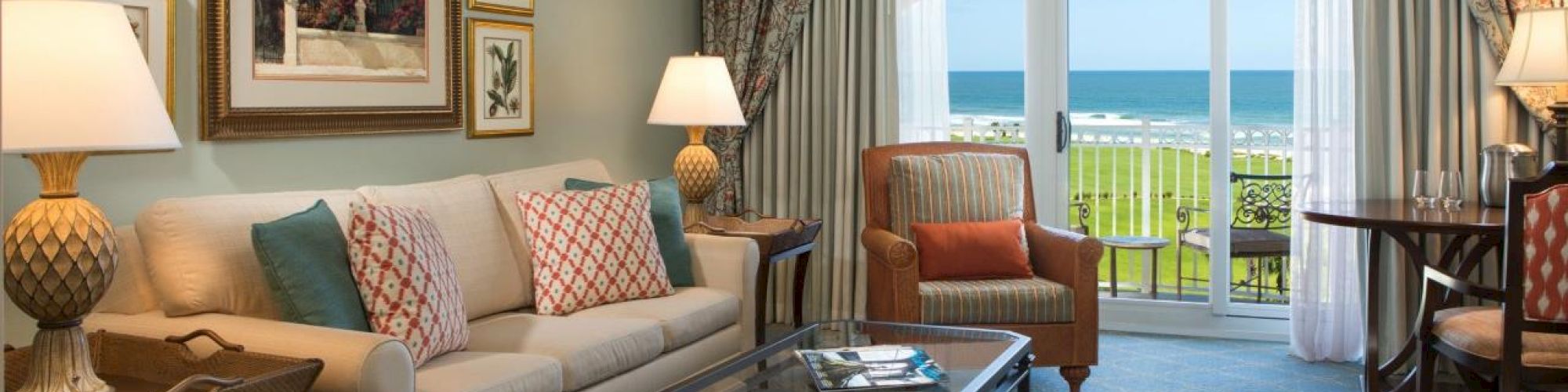 A cozy living room with a sofa, armchair, lamps, and framed art faces a balcony overlooking the sea, offering a relaxing view.