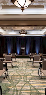 A spacious conference room set with rows of tables and chairs, two large projector screens, and a stage with blue lighting, ready for a presentation.