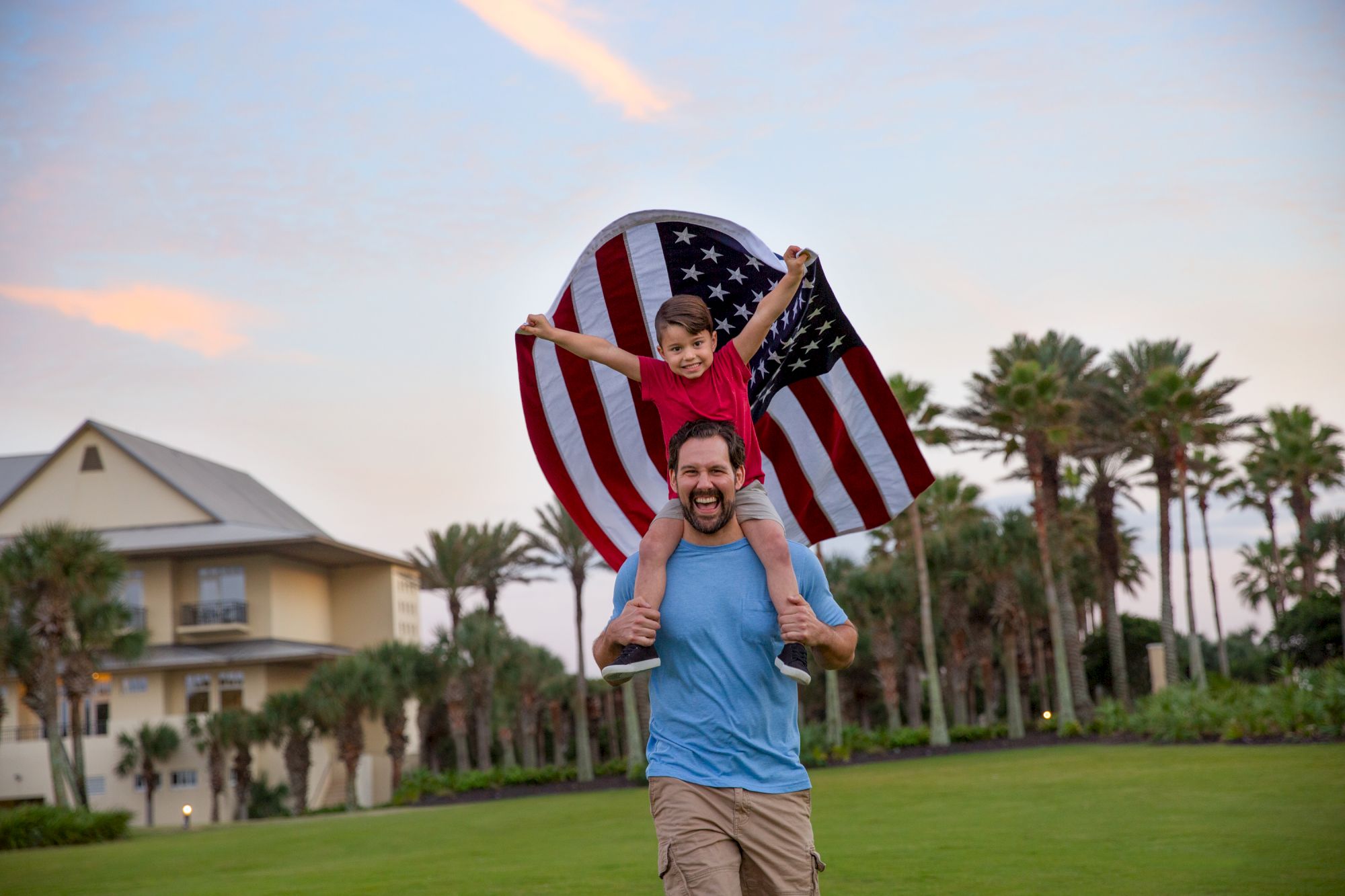 A man carries a happy child on his shoulders under a waving American flag in a sunny, palm-tree park.