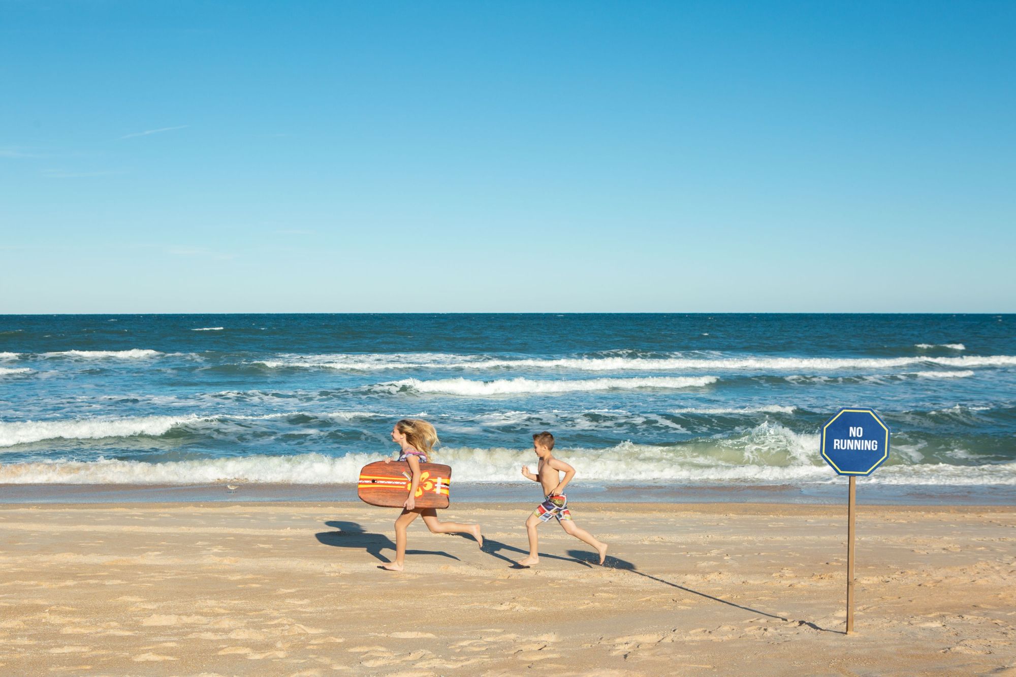 Two children running on a beach with a bodyboard, near waves. A sign reads "No Running."
