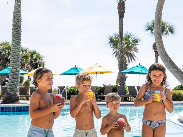 Four kids in swimwear stand in a pool, each holding a drink, with palm trees and blue umbrellas in the background, enjoying a sunny day.