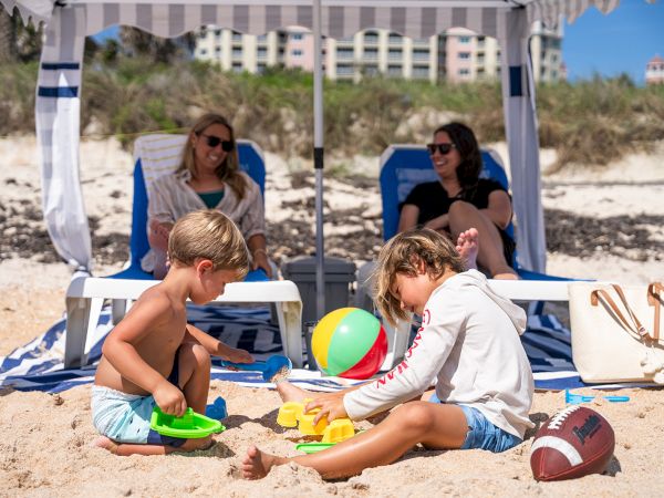 Kids playing in the sand under a striped canopy on the beach, with a ball and buckets; adults lounge nearby under the shade.