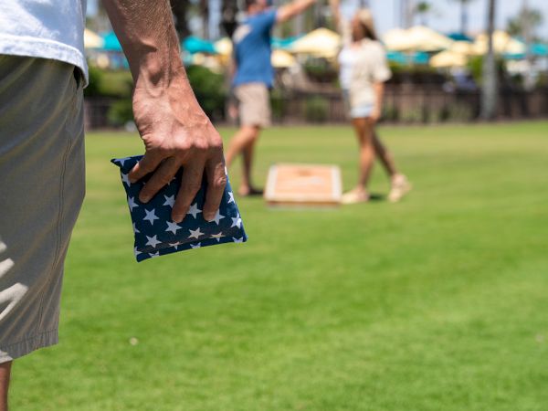 A man holds an American flag best-toy toss game area on a sunny lawn; foreground shows striped stars and stripes, with others playing in background.