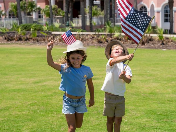 Two kids happily run on a sunny lawn, waving American flags with a pink-blue apartment building and palm trees in the background.