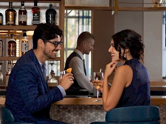 Two stylish people share drinks at a cozy bar, chatting and smiling as a bartender pours in the background.