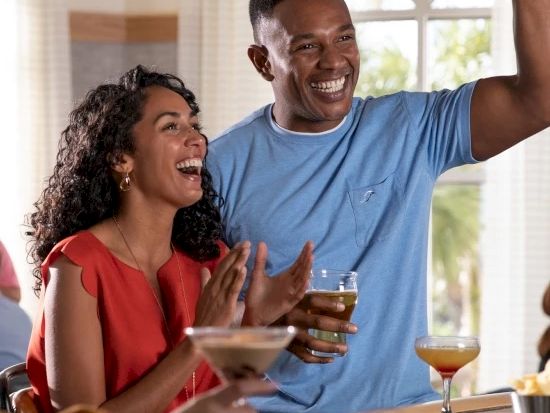 Two friends cheer at a bar or party, raising fists and clinking glasses, mid-laughter and celebration.