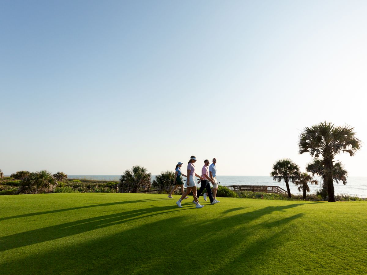 A group of people walking on a sunny, well-kept grassy field near the coast, with palm trees and long shadows at sunset or late afternoon.