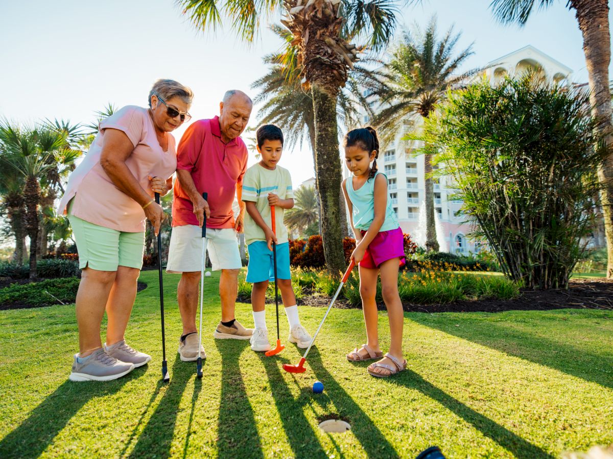 A family outdoors playing mini-golf on a sunny day, with two adults and two kids lining up shots on a green lawn near palm trees, smiling.