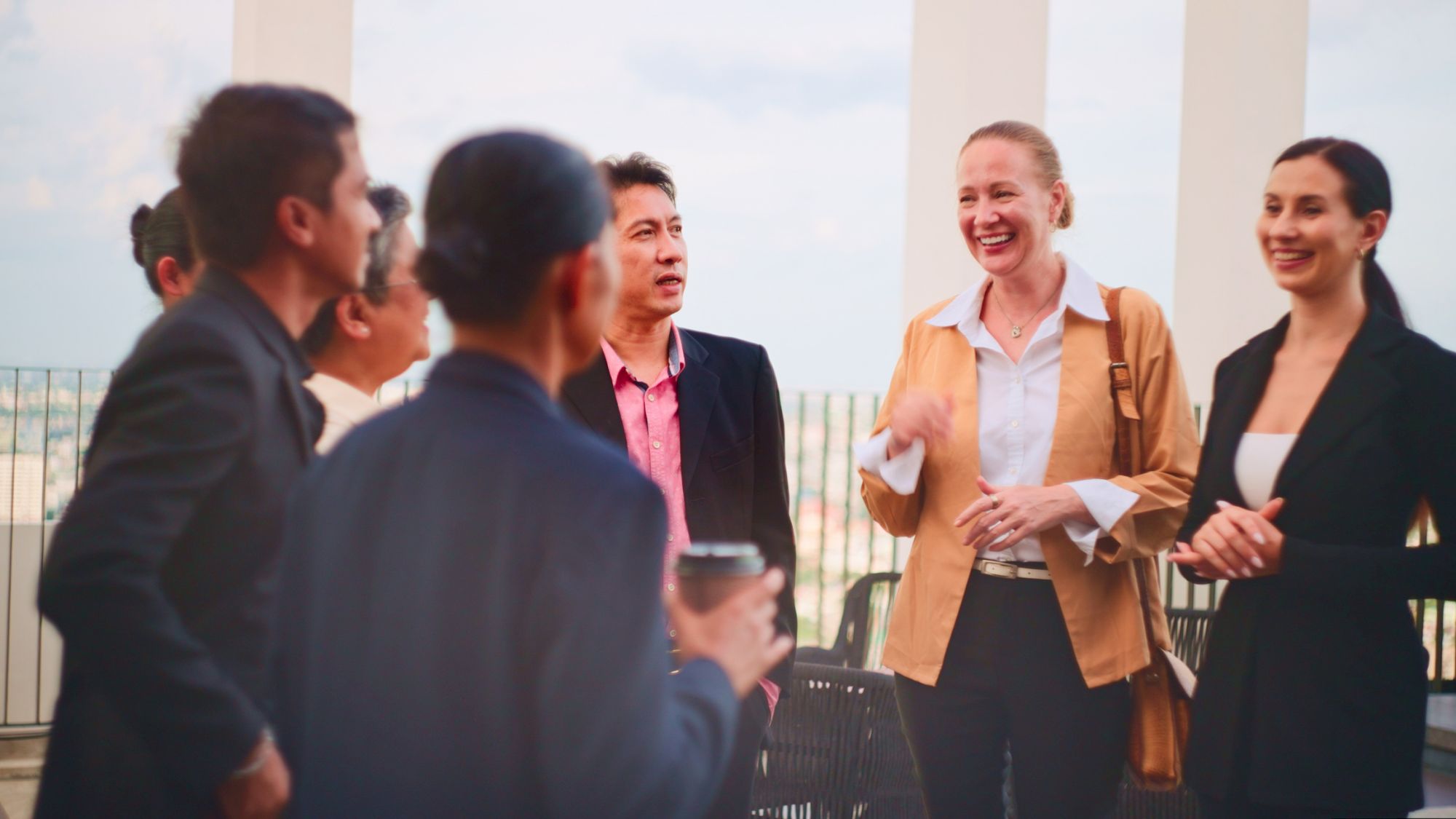 A group of professionals chat on a balcony, smiling and networking with drinks, dressed in business casual attire.