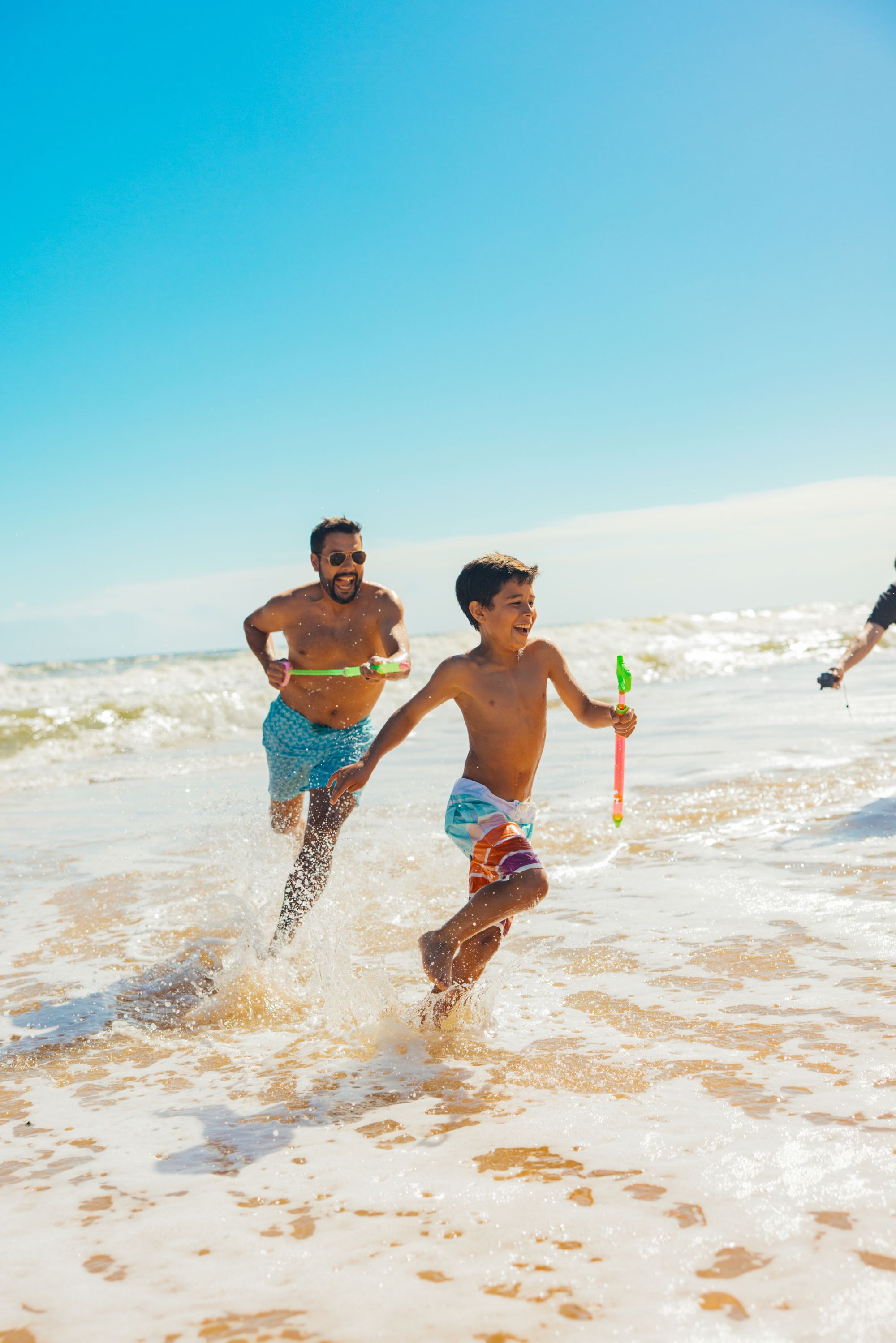 Two men run along the sunny beach, splashing through gentle waves; one holds a green toy while a blue sky and foamy shoreline frame their playful dash.