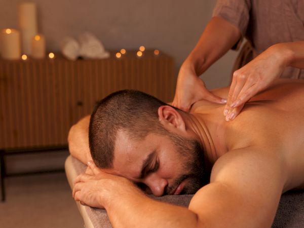 A man lies face down while receiving a shoulder/neck massage in a dimly lit room with candles in the background, relaxed and peaceful. end of sentence.