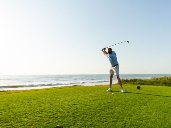 A golfer in a blue shirt swings on a sunny coastal golf course; the ball soars by the sea, with a tee and ball marker in the foreground.