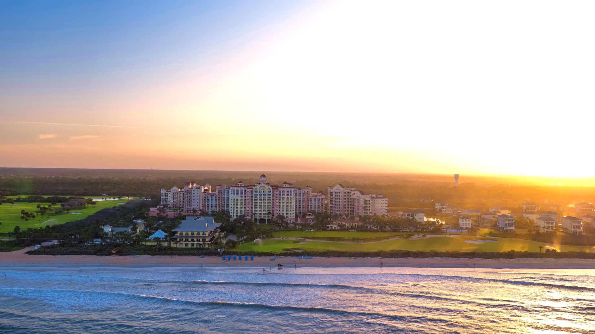 A coastal resort with buildings overlooks the ocean, set against a vibrant sunset or sunrise. Waves gently crash on the shore.
