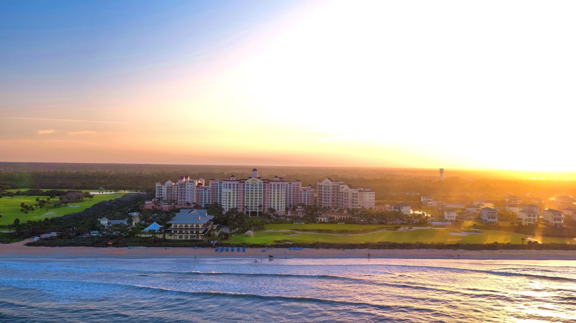 A coastal resort with buildings overlooks the ocean, set against a vibrant sunset or sunrise. Waves gently crash on the shore.