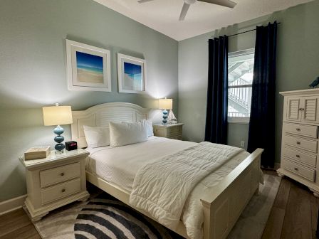 Cozy coastal bedroom with a white bed, matching nightstands, two lamps, coastal art, a dresser, and dark curtains by a window.
