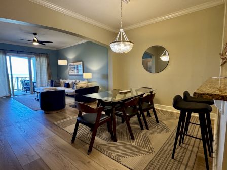 A modern open-concept living and dining area with a blue accent wall, a cozy seating area, a dining table, bar stools by a breakfast bar, and a sliding door to a balcony.