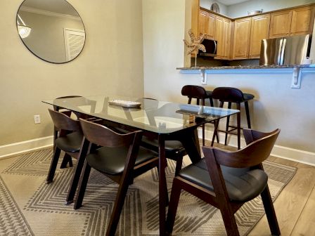 A modern dining area with a glass-top table and six dark wooden chairs, adjacent to a kitchen with wooden cabinets and bar stools, neat and inviting.
