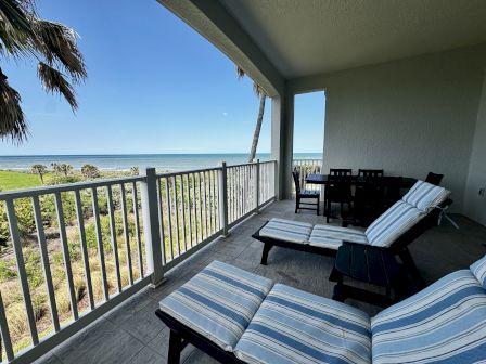 A beachfront balcony with striped lounge chairs, a dining table, and a view of the ocean and palm trees beyond.