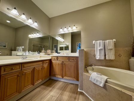A spacious bathroom with a double vanity, large mirrors, warm wooden cabinets, tiled countertops, and a separate bathtub with towels nearby, well-lit.