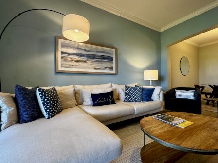 A cozy living room with a light beige sofa, blue and patterned cushions, a round wooden coffee table, wall art, and warm lighting.