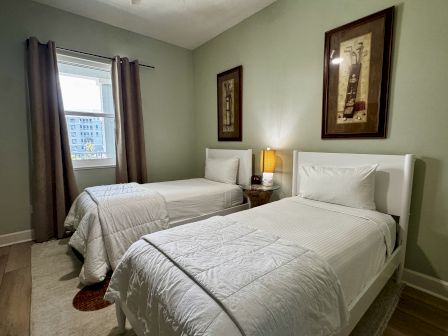 A tidy hotel room with two white-twin beds, a nightstand lamp, framed art, and a window with dark curtains.