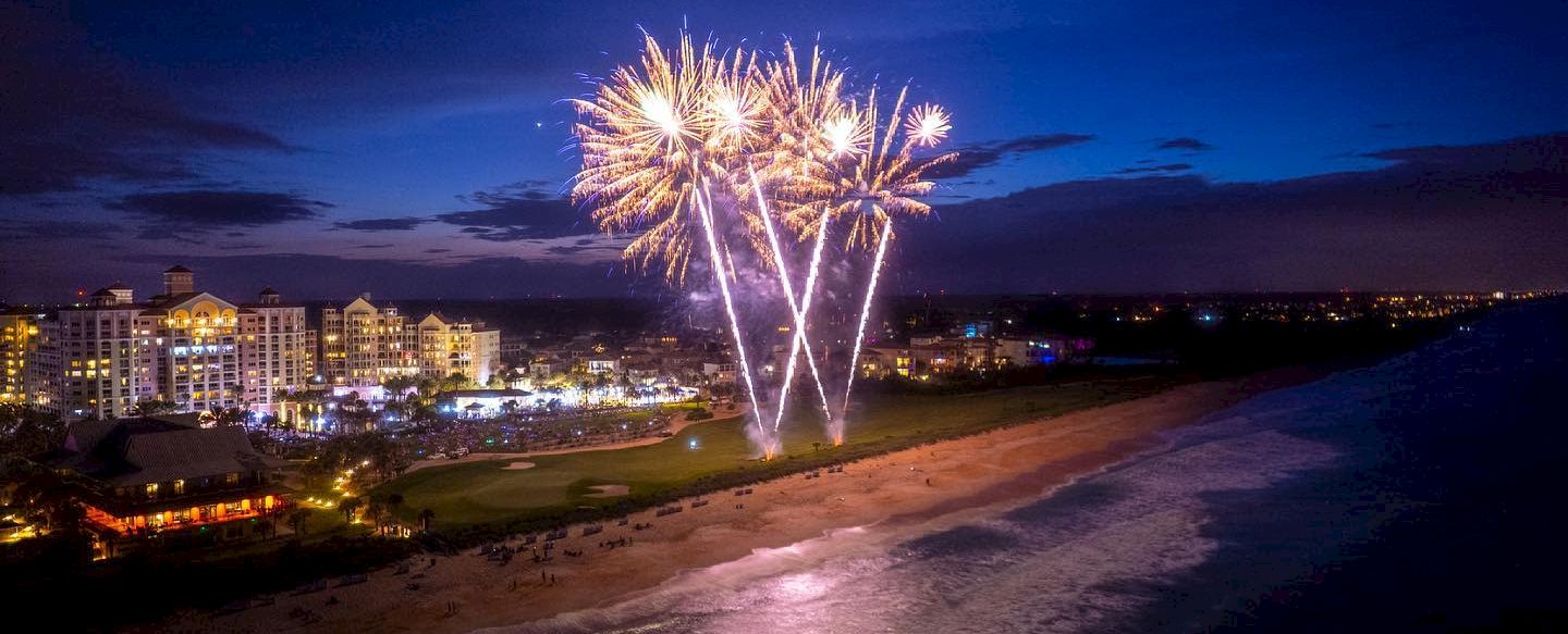 Fireworks burst over a coastal town at night, light reflecting on the waves; illuminated buildings line the shore as a beach glows below.