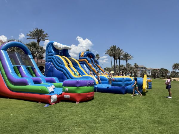 Colorful inflatable obstacle course set up on a grassy field, with kids playing and palm trees in the background under a sunny sky.