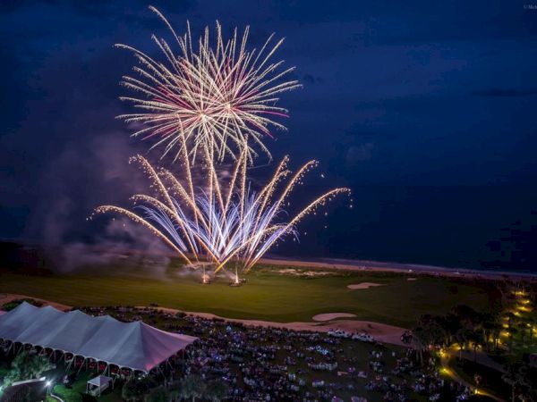 Fireworks bursting over a grassy field and beach at night, with a crowd and tents below, illuminating the sky with colorful streaks.