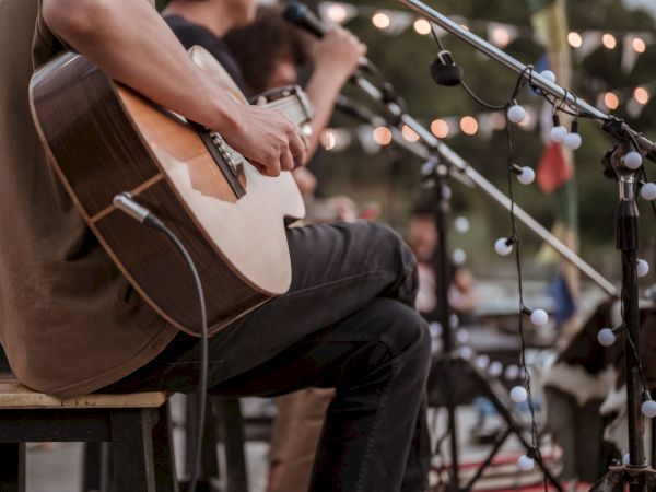 A guitarist strums an acoustic on a cozy outdoor stage, mic stands, string lights, and a small audience in the background.