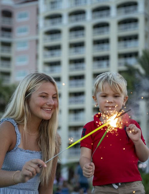 Two smiling people, a woman and a young boy, play with sparklers outdoors near a tall building and palm trees.