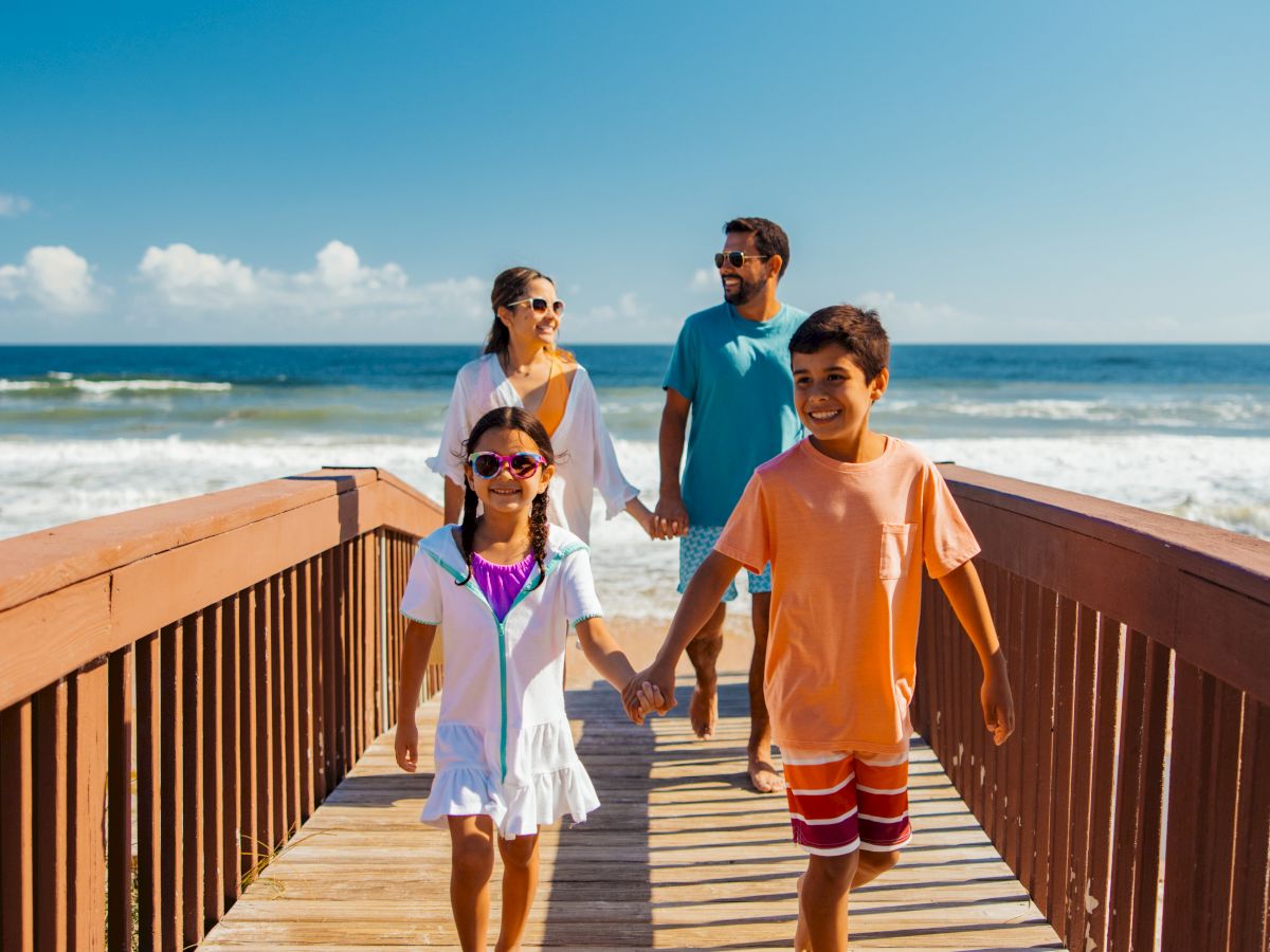 A family of four walks hand-in-hand on a wooden boardwalk toward the beach, bright sky, and ocean in the background.
