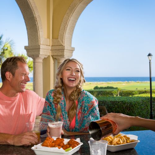 A couple enjoys a meal outdoors with drinks served, overlooking a scenic view of the sea and palm trees under an archway.