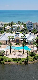 A seaside neighborhood with pastel houses surrounds a central circular pool area; water all around, with blue ocean beyond&mdash;paradise.