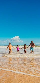 A family holds hands and wades through shallow ocean water on a sunny beach, smiling together as gentle waves roll in.