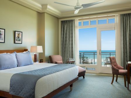 A bright hotel room with a king bed, many blue accents, a balcony door to an ocean view, and a small seating area with chairs and a desk.