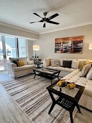 A modern living room with a sectional sofa, coffee table, lamps, wall art, and large windows.