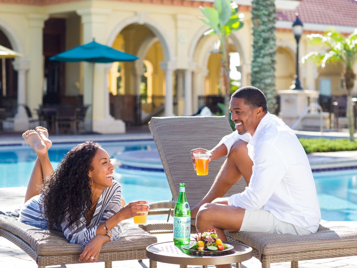 A couple lounges by a sunny pool, clinking drinks and smiling in a resort courtyard.