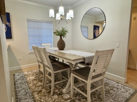 A light dining room with a distressed white table, six matching chairs, a large round mirror, a potted plant centerpiece, chandelier, and patterned rug.