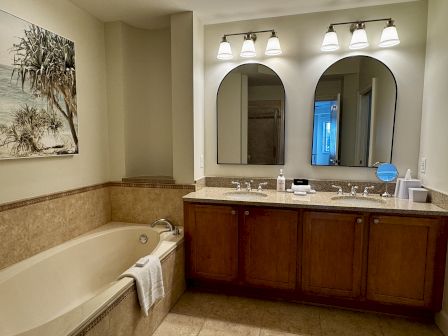 A modern bathroom with a tub to the left, double-sink vanity, large framed mirrors, warm wood cabinets, and soft overhead lighting.