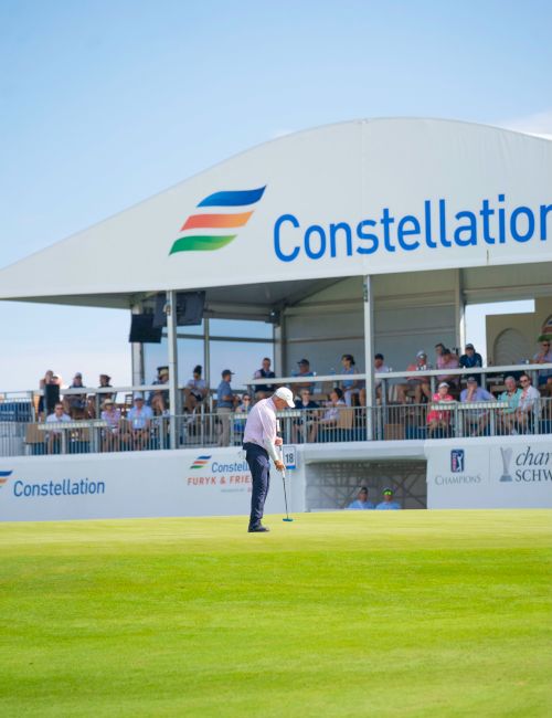 A golfer in a white cap practices/puts on a green while a &ldquo;Constellation&rdquo; tent and spectators sit behind.