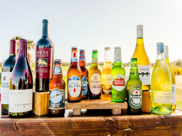 A lineup of assorted bottled drinks on a wooden table with a sunny outdoor backdrop, featuring wine, beer, and spirits in various colors.