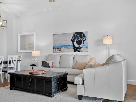 A bright living room with a white sectional sofa, black coffee table, lamps, and a wall painting of a dog by the seaside.