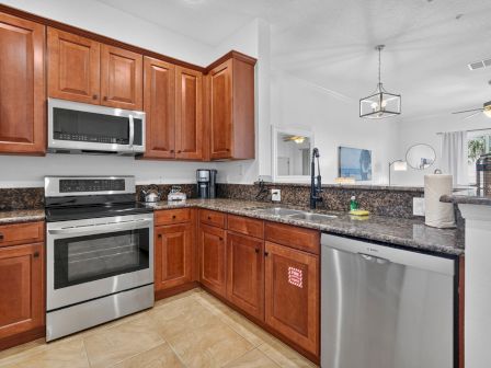 An empty modern kitchen with wooden cabinets, granite counters, stainless appliances, and a pendant light over the sink area.
