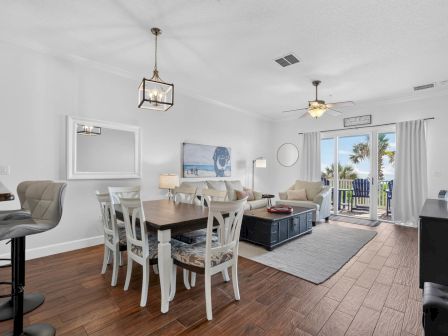 Bright beach-themed living room with white walls, dining table, sofas, ceiling fans, and palm trees visible through sliding doors.