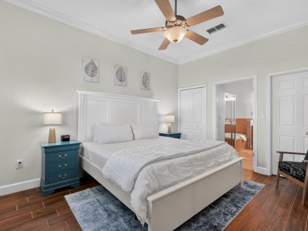 A bright bedroom with a white bed, blue nightstand, wall art, ceiling fan, and a door to an adjoining bathroom.