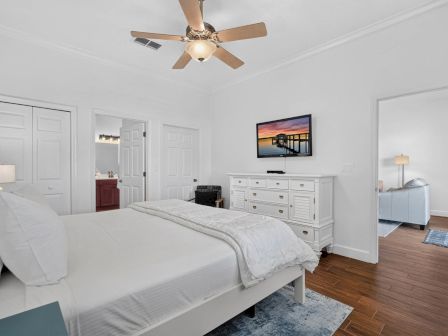 A bright bedroom with a white bed, ceiling fan, dresser with TV, and a doorway to a bathroom.