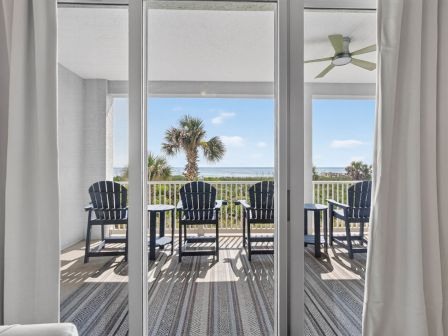 View from indoors to a seaside balcony with blue chairs, white railing, swaying palm trees, and sunshine in the distance.
