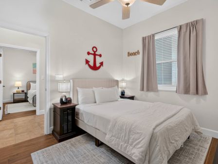 A bright bedroom with a white bed, gray curtains, bedside lamps, and a red anchor wall decal.