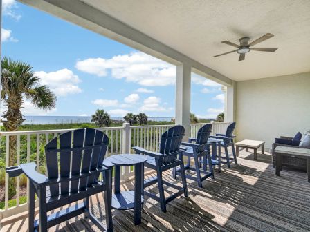 A seaside balcony patio with blue chairs, striped rug, a ceiling fan, and a view of the ocean and palm trees.